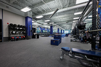 Weight room with REGUPOL aktivpro roll flooring in Thunder Gray, blue accents, and various workout equipment, including squat racks press, plyometric boxes and various other equipment.