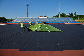 Workers unfold the turf onto REGUPOL abzorb at Spring Grove Area High School. A view of the high school stadium is located in the background.