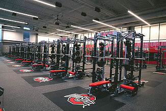 Ohio State University weight room with rows of weight racks and benches, bright overhead lights, and large windows. Featuring REGUPOL aktivpro roll flooring and crash zones inlaid with university logo.