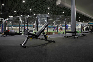 A inclined weight lifting bench rests on REGUPO aktiv rolled rubber flooring, with a view of the turf area in the background of Kelowna High Performance. There are a number of athletes located on the turf, receiving instruction from a coach. 