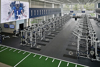 Overhead view of Penn State's football weight room with weight racks, benches, and turf area, showcasing the Penn State logo within REGUPOL aktivpro roll flooring and crash zones.