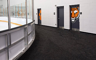 An angled view of the walkway around the Thayer Sports Center hockey rink, highlighting the curved shape of the rink and a doorway with the tiger logo leading into locker rooms. REGUPOL aktiv roll flooring was used throughout the walkway to ensure safety measures and durability are met for athletes.