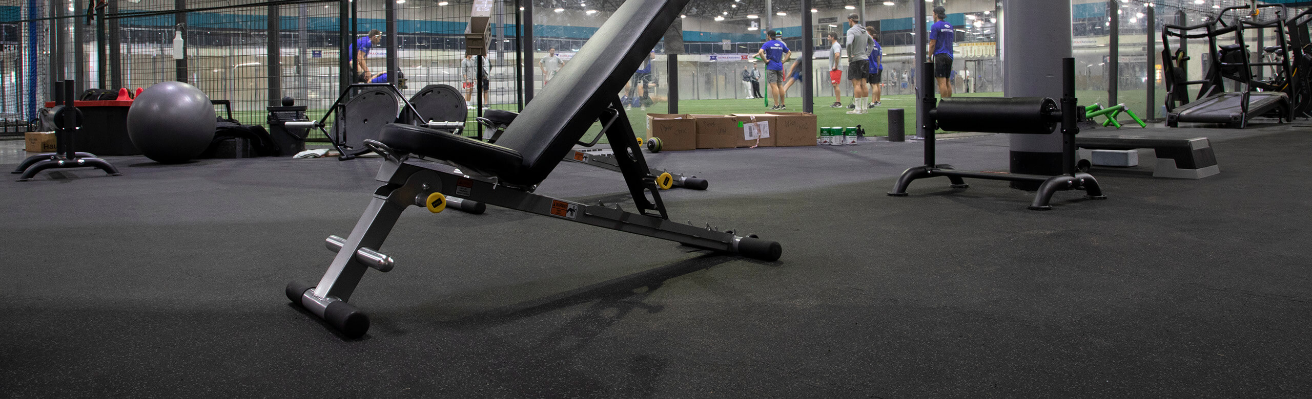 A inclined weight lifting bench rests on REGUPO aktiv rolled rubber flooring, with a view of the turf area in the background of Kelowna High Performance. There are a number of athletes located on the turf, receiving instruction from a coach. 