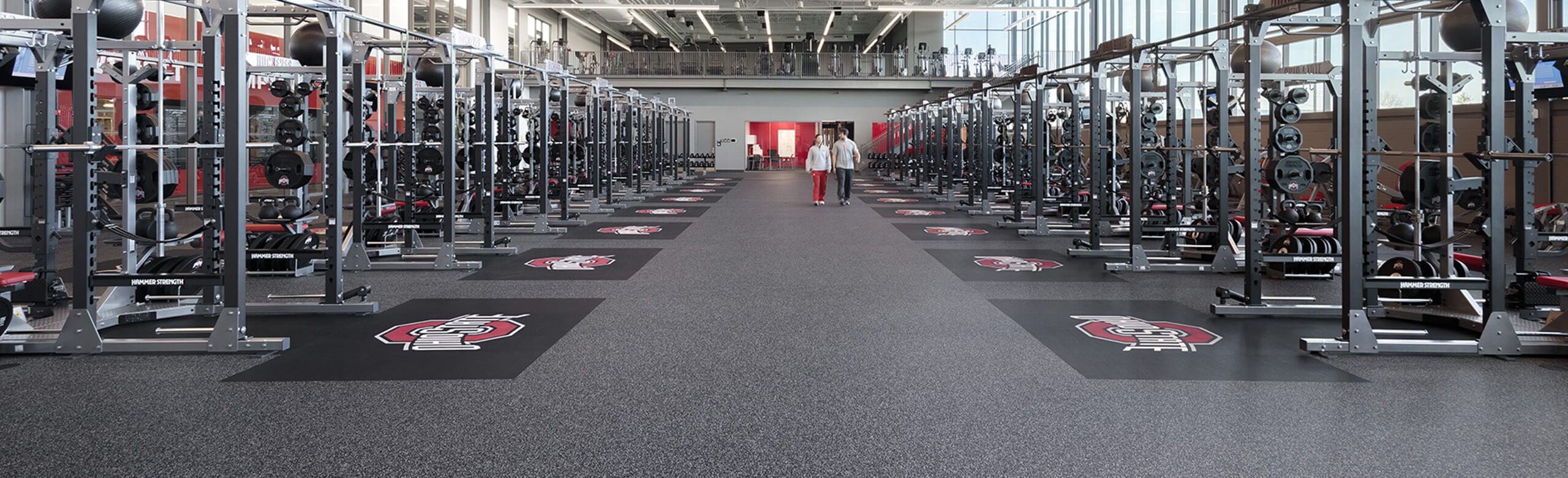 Ohio State University weight room with squat racks and weight benches, featuring the university's logo on a dark gray, shock-absorbing floor.