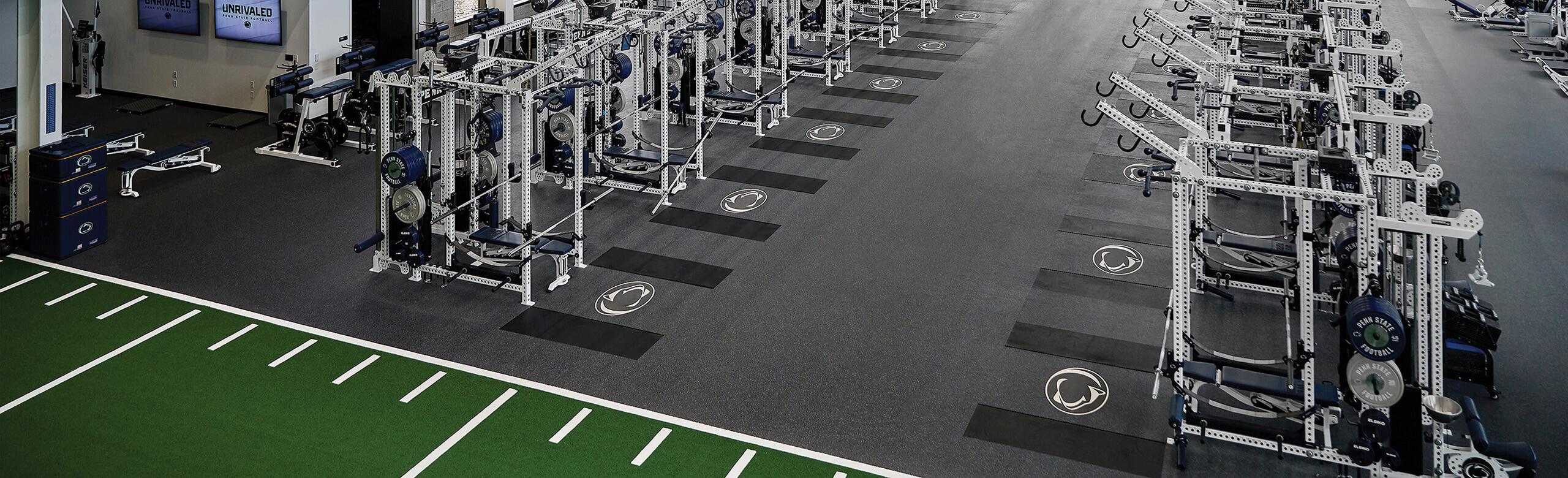 Overhead view of Penn State's football weight room with weight racks, benches, and turf area, showcasing the Penn State logo within REGUPOL aktivpro roll flooring and crash zones.